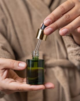 Female hands with natural manicure opening white green cosmetic dropper bottle. Close up of woman in bathroom wearing light brown bathrobe with drop of serum or essence falling from pipette