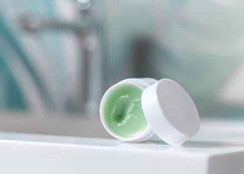 Opened white blank lip balm jar with a lid on washbasin with faucet in green tropical bathroom close up, packaging mockup. Lifestile scene with ubranded natural lipcare product