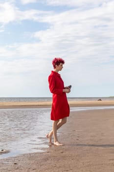 A woman walks barefoot on a sandy beach wearing a vibrant red dress. She gently holds a seashell while the calm sea reflects the cloudy sky behind her. The tranquil atmosphere creates a peaceful vibe.