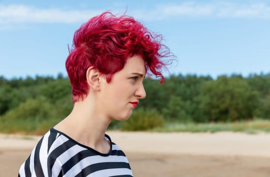 A woman with short, curly red hair walks along a sandy beach. She wears a striped shirt and looks thoughtfully into the distance, surrounded by lush greenery and a blue sky.