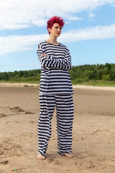 A young adult with curly red hair poses on a sandy beach, dressed in a loose striped outfit. The bright sky and surrounding trees add to the natural beauty of the setting.