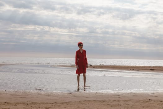A woman wearing a vivid red dress poses gracefully in shallow water near the shoreline. The sky is overcast, casting soft light on the scene.