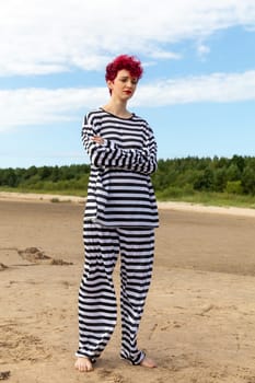 A person with vibrant red hair strikes a confident pose on a sandy beach. They wear a comfortable striped outfit, standing barefoot under a clear blue sky amidst lush green trees.