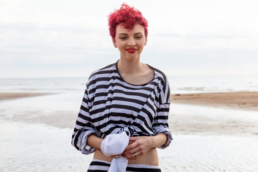 A woman stands on the beach with pink hair and a striped top, holding a towel. The shoreline features wet sand and gentle waves, with a cloudy sky in the background.
