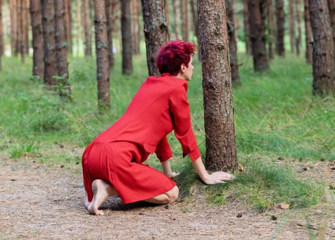A young woman with pink hair is kneeling in a lush pine forest, dressed in a vibrant red dress. She poses gracefully next to a tree, embodying beauty and tranquility in nature.