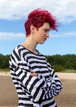 Individual with vibrant pink hair and a striped outfit stands outdoors, gazing thoughtfully at the landscape. The setting features a clear sky and lush greenery in the background.