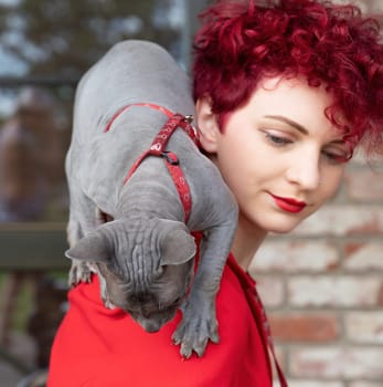 A woman with curly red hair and red lipstick carries a gray cat on her shoulder. They are outdoors, surrounded by a cozy atmosphere and natural light, enjoying a relaxed moment.