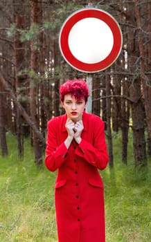 A woman with vibrant red hair poses in a striking red outfit in a forest. Behind her, a large no entry sign stands against the dense greenery, creating a contrast with her bold presence.