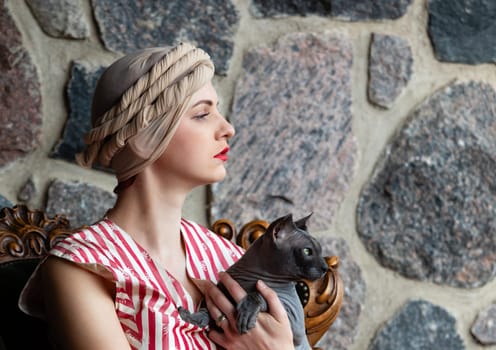 A stylish woman with a braided headscarf and red lips sits calmly, cradling a gray cat in her arms. The stone wall creates a rustic atmosphere, enhancing her vintage look.