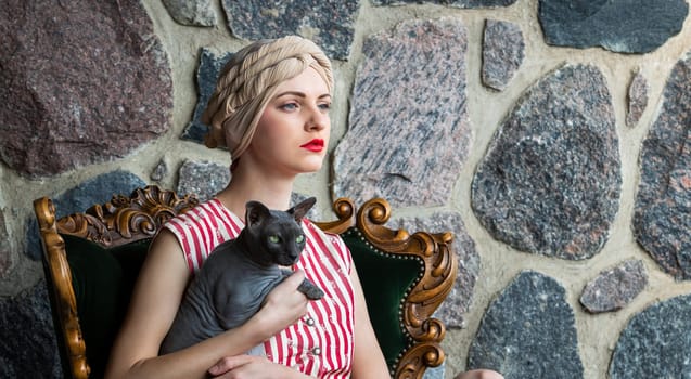 A woman with a head wrap sits calmly in an ornate armchair, holding a gray cat. The backdrop features a textured stone wall, creating a serene environment. Her expression is contemplative and poised.