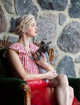 A woman in a fashionable dress and head wrap sits gracefully on an antique chair. She holds a cat closely, posing in front of a textured stone wall, creating an elegant atmosphere.