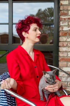 A woman with vibrant red hair wears a striking red outfit while sitting comfortably and holding a gray cat. The background features a bright, modern interior with large windows.