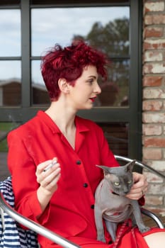 A woman with curly red hair sits comfortably on a chair, holding a grey cat in her lap. She wears a vibrant red outfit and enjoys a relaxed moment outdoors, surrounded by a tranquil setting.