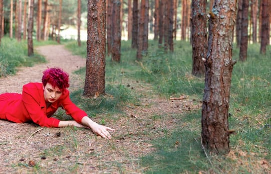 A young woman with pink hair wearing a red dress is lying on the ground in a serene pine forest. She embodies youth and beauty, surrounded by tall trees and soft green grass.