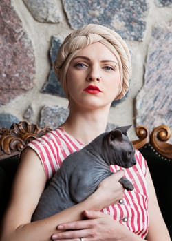 A woman with a stylish headwrap and striking red lips holds a sphynx cat close. She poses confidently against a rustic stone wall, exuding vintage charm and elegance.