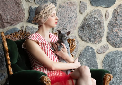 A woman dressed in vintage fashion sits gracefully in a plush chair, cradling a cat close to her chest, with a backdrop of textured stone walls. The soft lighting enhances the serene atmosphere.