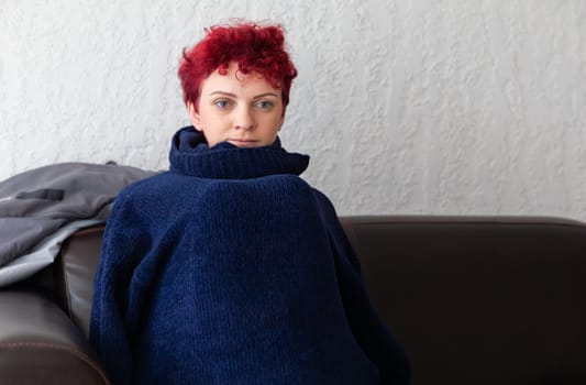 A person with curly red hair sits comfortably on a dark brown couch, wrapped in a large navy sweater. The light and textured wall adds a cozy feel to the indoor environment.