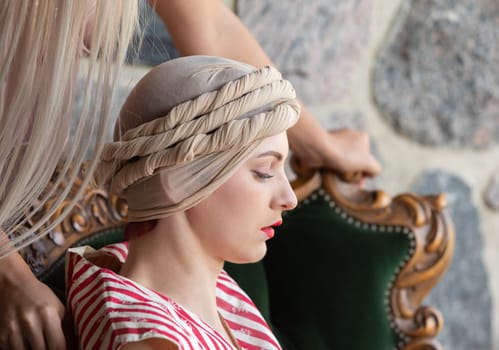 A woman with a refined look sits gracefully on a vintage chair. A hairstylist carefully works on her hair, creating an intricate updo.