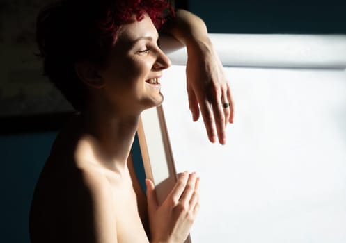 A young woman with short, curly hair is smiling while leaning against a blank canvas. Soft light illuminates the room as she engages in her art session, immersed in creativity.