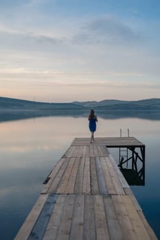 A young woman with wet hair, wrapped in a blue towel, sits on a wooden dock by a calm lake during sunset.
