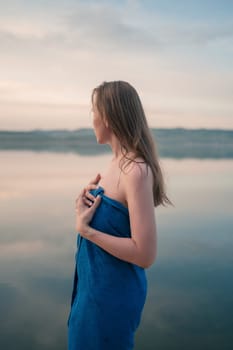 A young woman with wet hair wrapped in a blue towel stands near a calm lake at sunset, gazing thoughtfully into the distance.