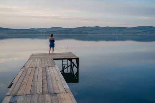 A young woman with wet hair, wrapped in a blue towel, sits on a wooden dock by a calm lake during sunset.