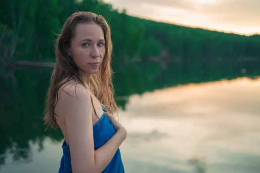 A young woman with wet hair wrapped in a blue towel stands near a calm lake at sunset, gazing thoughtfully into the distance.