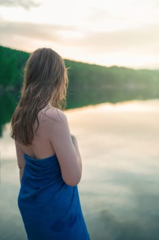 A young woman with wet hair wrapped in a blue towel stands near a calm lake at sunset, gazing thoughtfully into the distance.