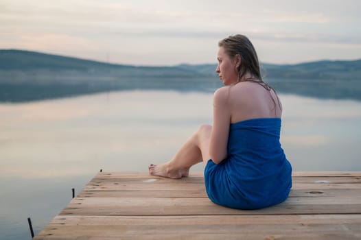 A young woman with wet hair, wrapped in a blue towel, sits on a wooden dock by a calm lake during sunset.