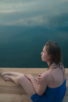 A young woman with wet hair, wrapped in a blue towel, sits on a wooden dock by a calm lake during sunset.