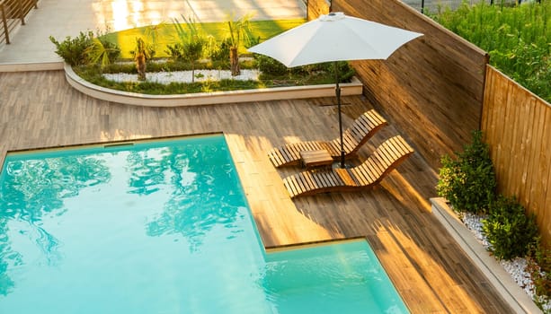 Luxurious poolside wooden deck with curved lounge chairs, umbrella, and turquoise swimming pool in a sunny backyard