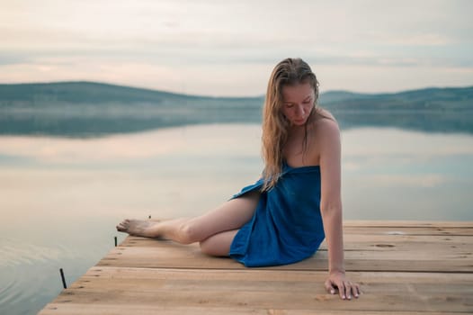 A young woman with wet hair, wrapped in a blue towel, sits on a wooden dock by a calm lake during sunset.