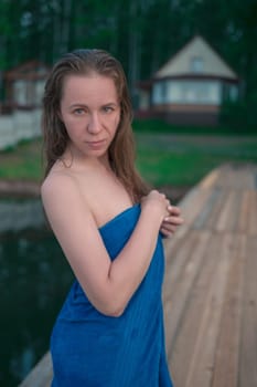 A young woman with wet hair wrapped in a blue towel stands near a calm lake at sunset, gazing thoughtfully into the distance.