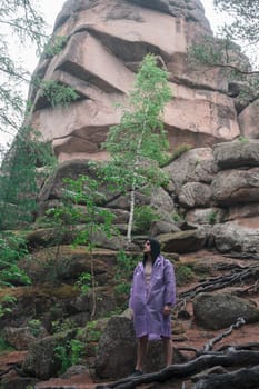 A man wearing a purple raincoat stands among rugged rocks and towering formations surrounded by lush forest greenery. The Stolby nature reserve park, Krasnoyarsk