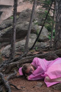 A woman in a flowing pink dress sleeps on the ground under twisted tree roots in a forest with towering rock formations.