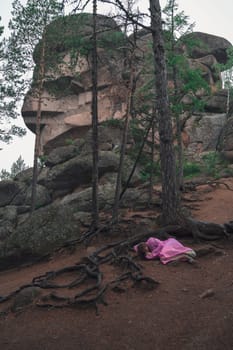A woman in a flowing pink dress sleeps on the ground under twisted tree roots in a forest with towering rock formations.