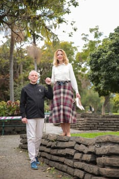 Couple holding hands and walking in a park, the woman balancing barefoot on a stone wall