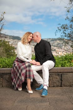 Happy mature couple holding hands and touching foreheads while sitting on a stone wall, enjoying the panoramic view of funchal