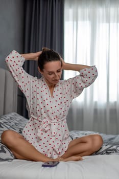Woman in a white robe sits cross-legged on a bed, hands behind her head, near a remote control. Morning routine in a bedroom.