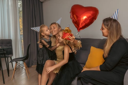 A woman in a gold dress receives kisses and a gift from two children wearing party hats, while another woman watches a red heart balloon floats nearby. Happy birthday celebration.