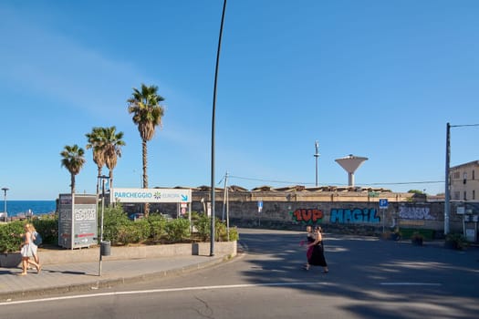 Catania. Italy - February 25, 2025: A sunny day at a coastal parking area in Catania with palm trees, graffiti on the walls, and people walking. Concept: Urban landscape