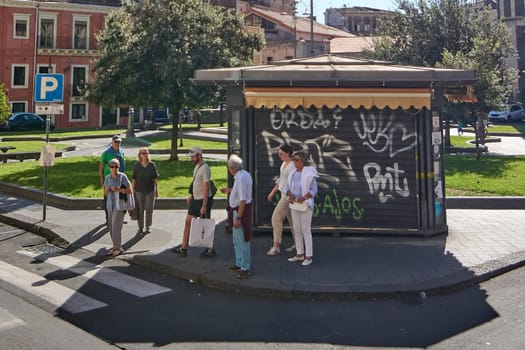 Catania, Italy - February 25, 2025: Tourists and locals gather at a graffiti-covered bus stop in Catania, Sicily, under sunny skies, near a park. Concept: Urban daily life