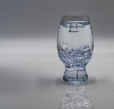 A clear glass filled with water and ice cubes sits on a smooth, reflective surface. The light highlights the clarity of the liquid and the ice, creating a refreshing appearance.