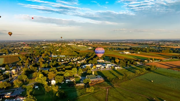 Hot air balloons of various colors drift gracefully over a scenic countryside at sunrise. The serene landscape features fields, trees, and quaint houses, creating a beautiful morning view.