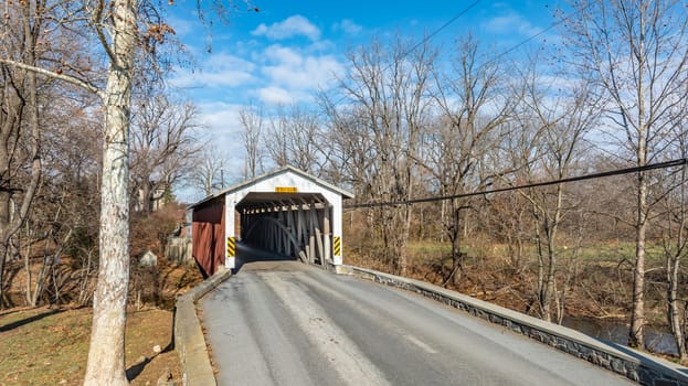 A charming covered bridge spans a tranquil road in a rural setting. The sunny winter day highlights the bare trees, making this scene picturesque and serene.