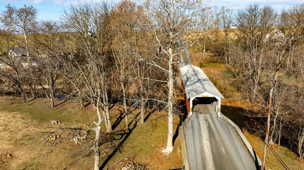 A historic covered bridge spans a quiet road in a rural area, surrounded by bare trees and colorful autumn foliage. The scene reflects a calm and picturesque landscape.