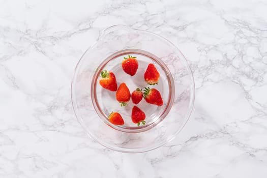 Ripe strawberries are submerged in water within a large glass mixing bowl, a step in washing the fruit to ensure cleanliness and longevity before storage or consumption.