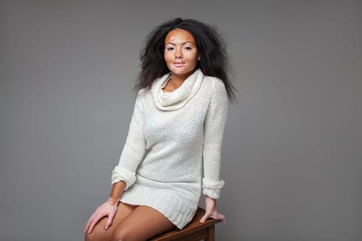 Confident female fashion model with vitiligo skin disease sitting on stool against gray background. Portrait of young lady is wearing white turtleneck sweater mini dress in studio