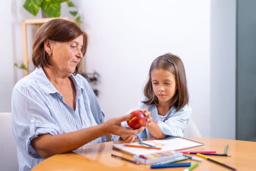 Grandmother placing apple in girl's hand as she sits at table with colored pencils and book, creating warm and nurturing moment filled with creativity and care