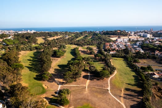 Aerial drone view captures expansive golf course in Estoril, Portugal, with its meticulously manicured greens, tree-lined fairways, and scenic surroundings